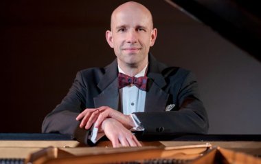 Christopher Taylor in a suit leaning on his piano looking at the camera. 
