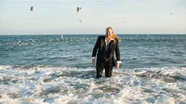 A man wearing a suit stands in the ocean with seagulls flying in the background.