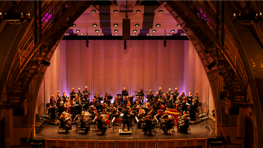 A chamber orchestra and a choir perform onstage in warm lighting.
