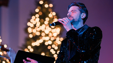 A man wearing a black sparkly blazer sings into a microphone with lights in the background.