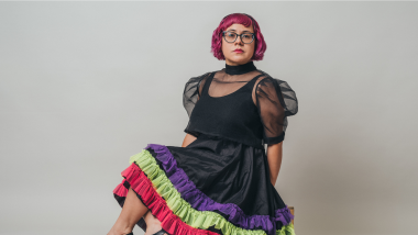 A woman with short, hot pink hair and glasses poses, seated in a black dress with a colorful trim.