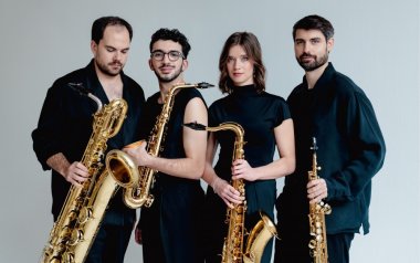Arcis Saxophone Quartet poses in all black outfits with their instruments in front of a blank wall.
