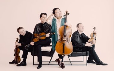 The four members of the Calidore String Quartet sit on seats together looking off to the side while holding their string instruments.