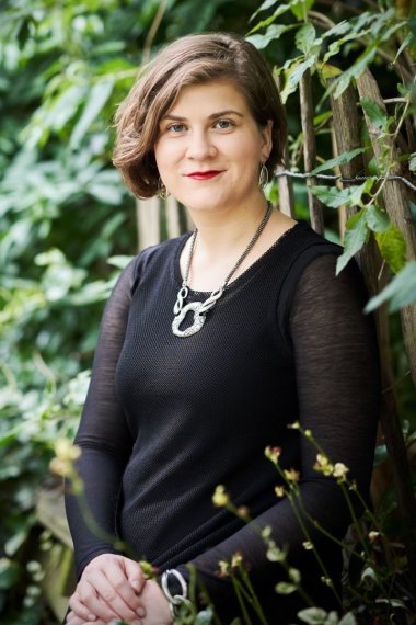 A women in black formal attire stands in front of greenery. 