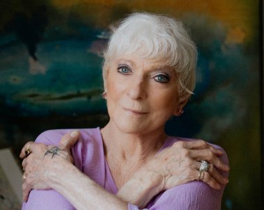 A women with short white hair in a violet colored attire standing in front of a turquoise watercolor backsplash.