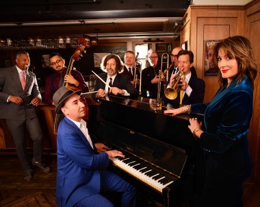 The band is standing around the piano posing in a cozy backdrop