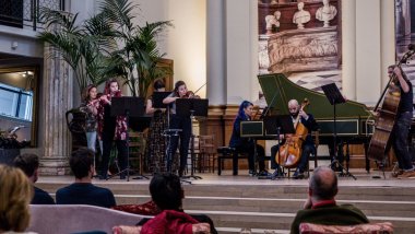 musicians playing flute, violins, harpsichord, cello and bass on a classical style stage with palms
