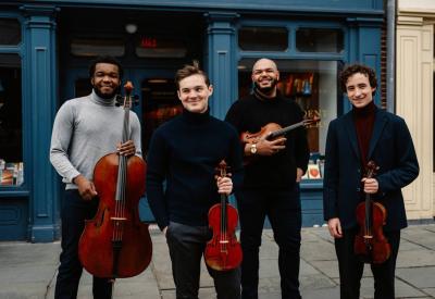 The Isidore String Quartet stands in front of a blue building, each holding their instruments