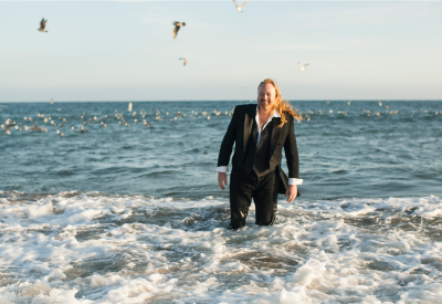 A man wearing a suit stands in the ocean with seagulls flying in the background.