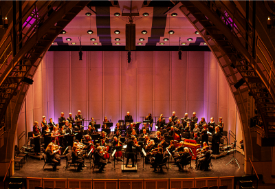 A chamber orchestra and a choir perform onstage in warm lighting.