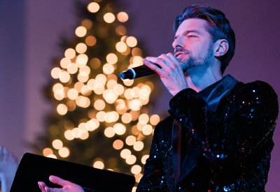 A man wearing a black sparkly blazer sings into a microphone with lights in the background.