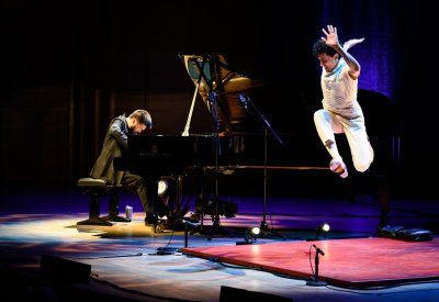 A man plays piano on stage while a male dancer leaps in the air next to him.