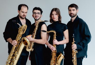 Arcis Saxophone Quartet poses in all black outfits with their instruments in front of a blank wall.