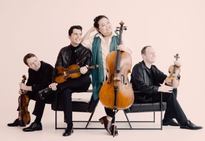 The four members of the Calidore String Quartet sit on seats together looking off to the side while holding their string instruments.