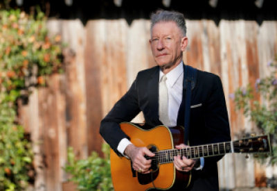 Lyle Lovett in a suit, with holing a guitar, posing in front of a brown, tan, and faded wooden fence.