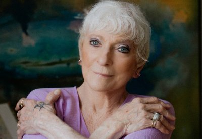 A women with short white hair in a violet colored attire standing in front of a turquoise watercolor backsplash.