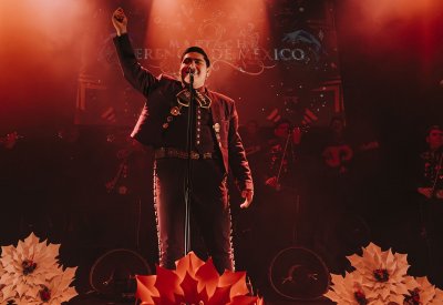 A man is dressed in traditional a Mexican mariachi attire standing in front of a mic with his hands up. There are flow appliqués at the bottom of the frame. 